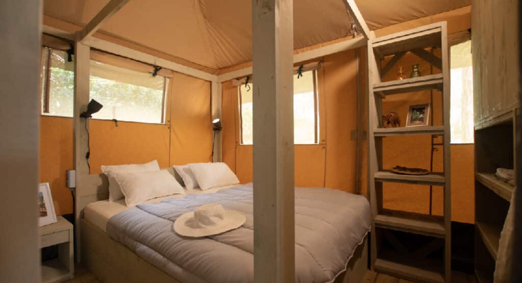 Cozy bedroom inside a safari tent at Yelloh! Village Sant Miquel campsite. The room features a comfortable double bed with white pillows and a light grey duvet, accented by a sun hat. Soft lighting and large windows create a warm, inviting atmosphere. A wooden shelf on the side holds decorative items, adding to the room's rustic charm.