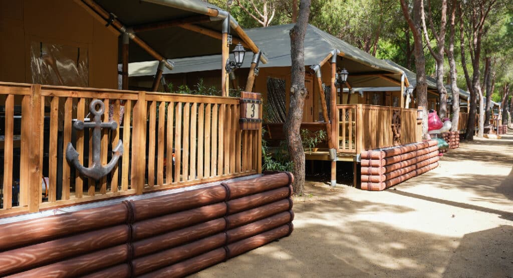 Exterior view of safari tents at Yelloh! Village Sant Miquel campsite. The tents are set on wooden platforms with log-style railings and feature nautical-themed decorations, such as an anchor on the fence. The area is shaded by tall pine trees, creating a cozy and natural atmosphere along a sandy pathway. The tents are well-spaced, providing a comfortable and inviting environment for guests.